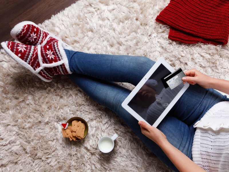 Woman holding a digital tablet and a credit card in seasonal attire on a shag rug with milk and Christmas cookies.