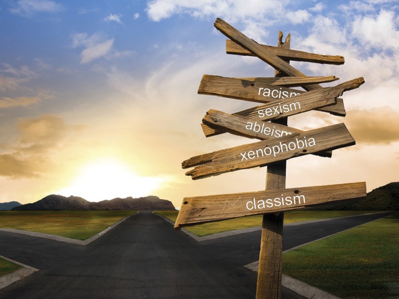A visual representation of the term intersectionality with a wooden sign placed at a crossroads pointing the way towards racism, sexism, ableism, xenophobia and classism.