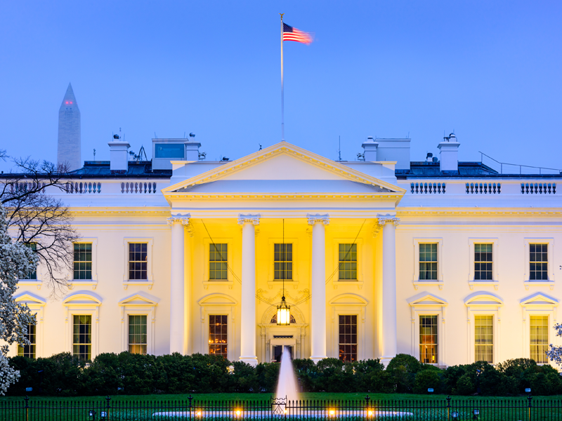 Image of the front of the White House at nighttime with an American flag flying proudly upon the rooftop.