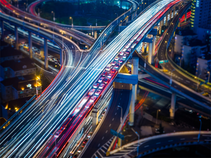 Long exposure photo of cars on a highway overpass. The left lane is a blur of white showing the traffic moving while the right lane is a dotted canvas of tail lights showing a major traffic jam. Soft blues from a twilight sky mix with the rough orange of urban life. Streetlights in the background reveal a world underneath the overpass one that is dark both metaphorically and literally. The photo shows concrete life coming to fruition. The buildings, the overpass itself, all of the same material. What is this urban life? It is a complex mix of blue and orange on a concrete palette drowning out the cries of a lonely green park. Just a hint of the once expansive natural wonder of this land exists surrounded by the overpass, another metaphor. The overpass is layered much like the way we experience life. While we may wait trapped in traffic above, below the cars race to and fro, trapped in an inevitable push for space. Amidst the chaos of urban life a red light frees lanes in the bottom right. Show the constructed nature of this problem we call traffic. By mans design traffic comes and goes, ebbing and flowing not from the tides but from the red lights that dot the image.